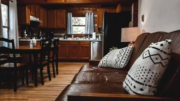 Een gezellige woonkamer met een leren bank, gestreepte kussens, een eethoek met houten stoelen, en een keuken met houten kastjes en een koelkast.