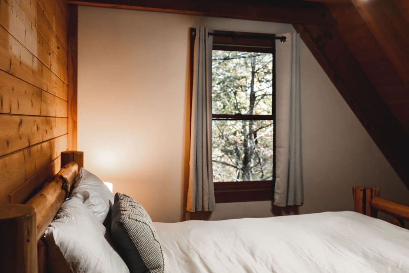 Un lit simple avec une tête de lit en bois, dans une pièce aux murs en bois et dotée d’une fenêtre avec des rideaux.