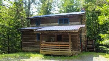 Une cabane en bois rustique, dotée d’un porche et d’une cheminée en pierre, entourée d’une forêt dense.