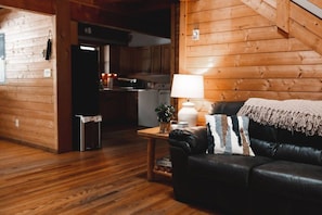A cozy living room with a wooden floor, a black leather sofa, a lamp, and a kitchen in the background. - Wolf Creek - Hot Tub/No Pets (Purlear)