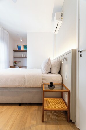 A neatly arranged hotel room with a bed, a bedside table with a cup, a wall-mounted air conditioner, and a small shelf with decorative items. - Equalize Vila Mariana by Viva (São Paulo)