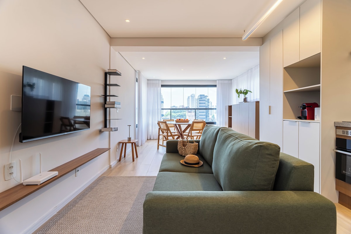 A modern living room with a flat-screen TV, a green sofa, a dining area with wooden chairs, and a city view through large windows.