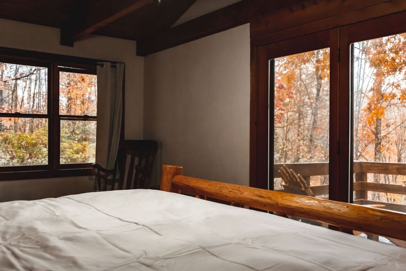 A room with a bed, a chair, and large windows with a view of autumn trees.
