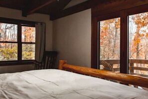 Une chambre avec un lit, une chaise et de grandes fenêtres donnant sur les arbres aux couleurs automnales.