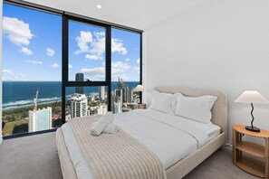 A bedroom with a large bed, two bedside lamps, and a view of the city and ocean. - Elegant 2BR with Panoramic Water Views (Broadbeach)