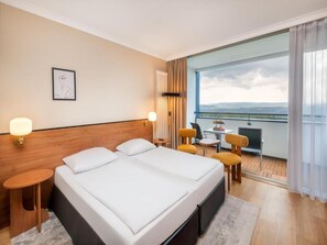 Ein Hotelzimmer mit einem großen Bett, einem kleinen Tisch, zwei Stühlen, einem Balkon mit Aussicht und einem gerahmten Bild an der Wand.