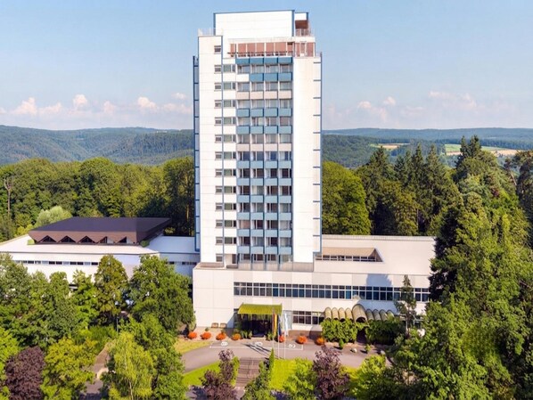 Ein modernes Hotel mit einem zentralen Turm, umgeben von üppiger Vegetation und einer Bergkulisse.