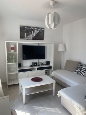 Un salon moderne avec un téléviseur à écran plat, des meubles blancs, une assiette décorative rouge posée sur une table basse blanche, et un tableau représentant un paysage côtier.