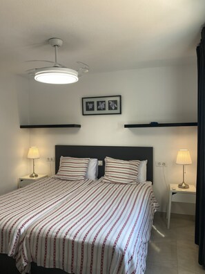Une chambre d’hôtel comprenant un lit, deux tables de chevet, un ventilateur de plafond, un tableau accroché au mur et une petite table de nuit avec une lampe.