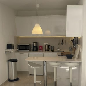 Une cuisine moderne avec des meubles blancs, un évier en acier inoxydable, un four à micro-ondes, une machine à café et un luminaire suspendu.
