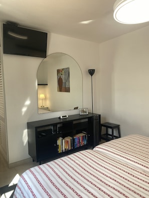 Une chambre à coucher avec un lit, un téléviseur fixé au mur, un bureau avec une lampe, une bibliothèque et un miroir.