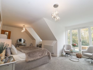 Ein Schlafzimmer mit schrägverdeckter Decke, einem Bett mit Felldecke, einem Schminktisch, einem Spiegel, einem Heizkörper, einem Sessel, einem Tisch mit einer Vase und einem Fenster mit Blick auf Bäume.