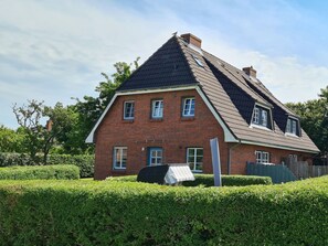 Ein zweistöckiges Backsteinhhaus mit einem steilen Dach, zahlreichen Fenstern und einer überdachten Sitzgelegenheit im Freien.