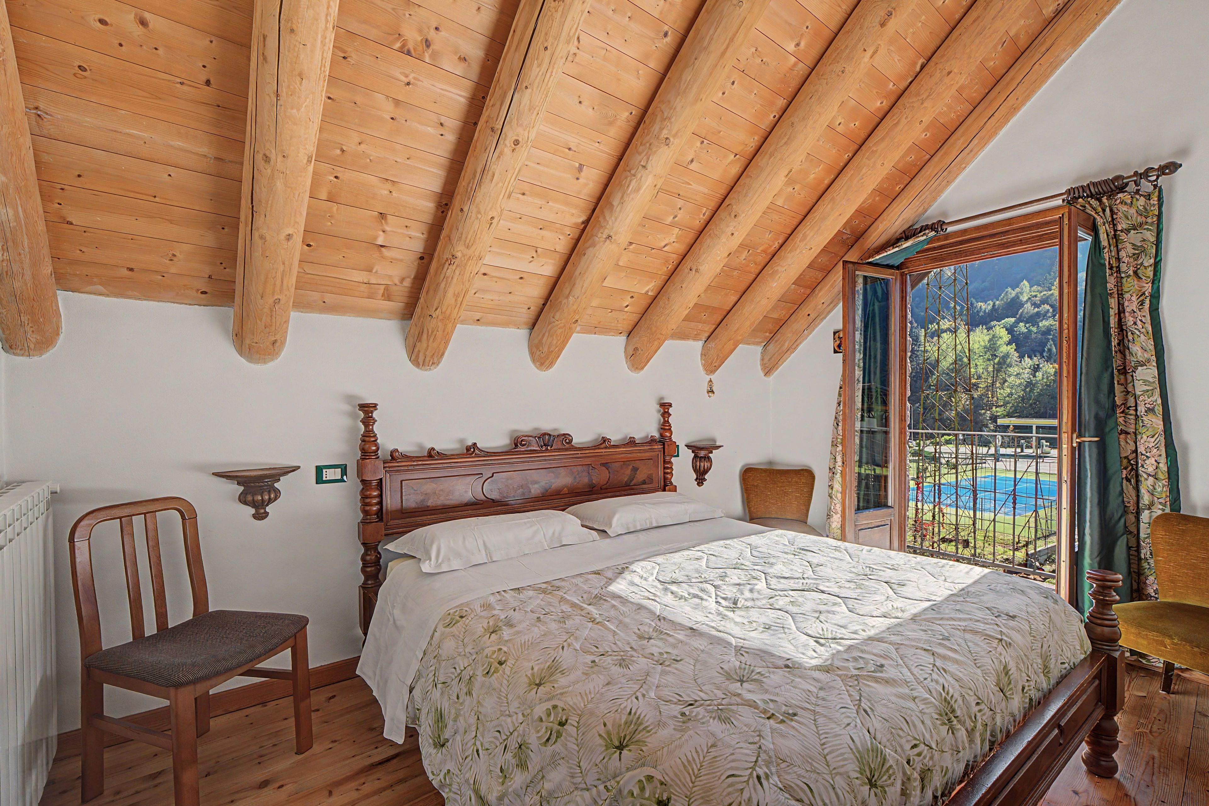 A bedroom with a wooden ceiling, a bed with a floral bedspread, a chair, and a window with a view of a pool and mountains.