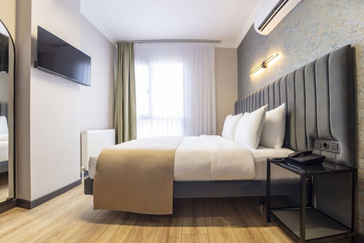 A modern hotel room with a large bed, a bedside table with a phone, a flat-screen TV mounted on the wall, and a window with curtains.