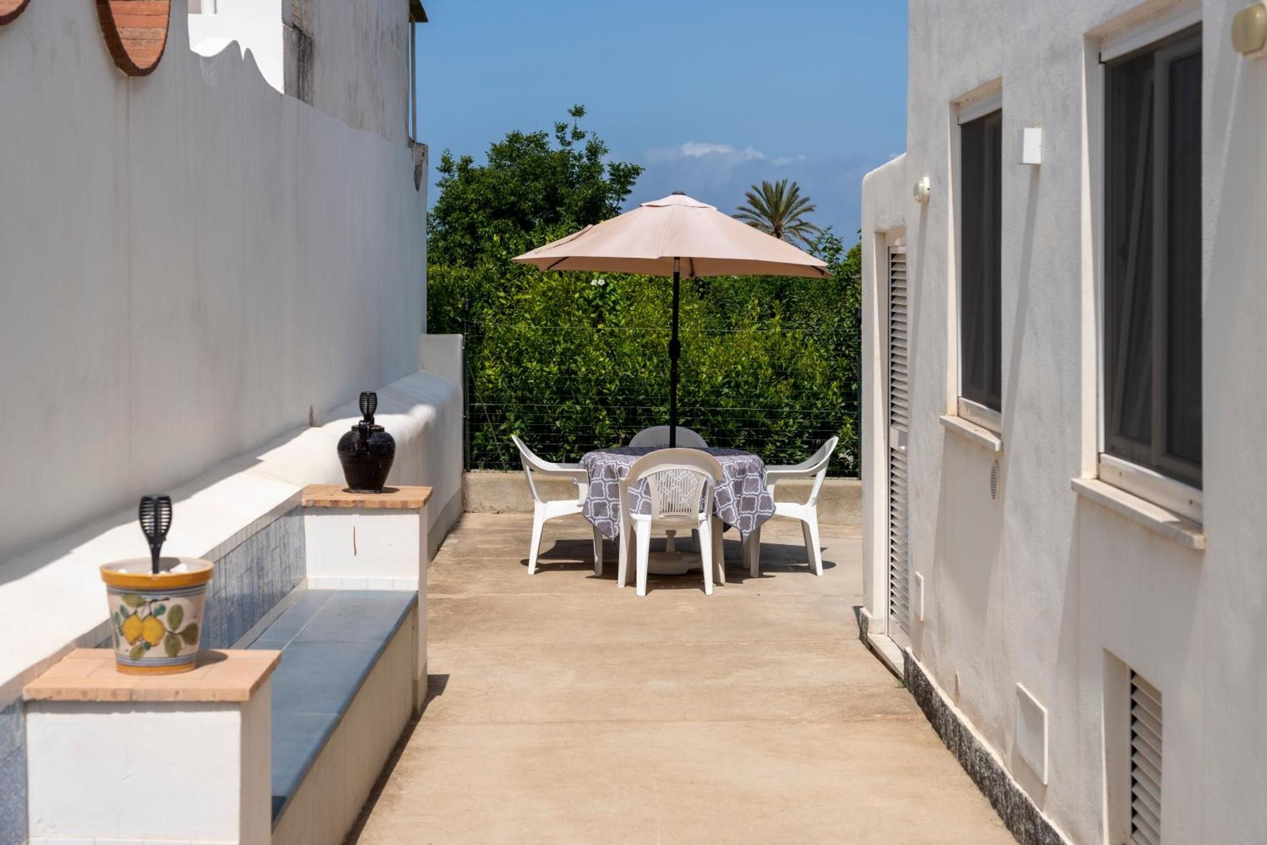 Un estrecho pasillo exterior con una mesa y sillas bajo un paraguas grande, una planta en maceta y un jarrón decorativo.