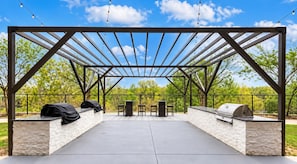 Un patio exterior con una estructura de pérgola, dos parrillas de barbacoa y una zona de estar con mesas y sillas.