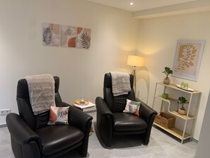 A room with two black leather chairs, a small side table with a candle, a floor lamp, and a shelf with decorative items.