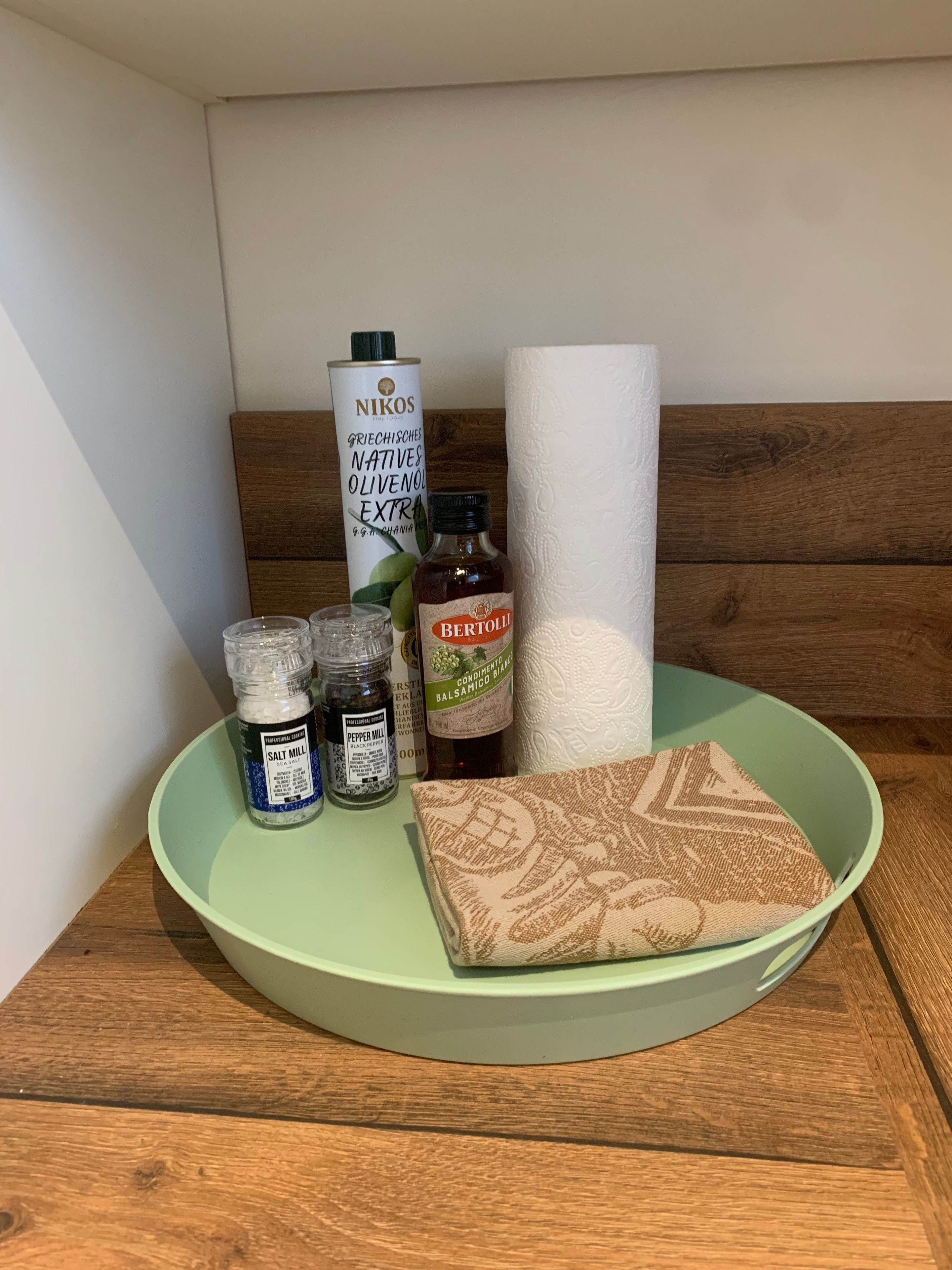 Een groene dienblad met diverse items, waaronder een fles BERTOLLI balsamico azijn, een zoutstrooier, een pepermolen en een rol keukenpapier.