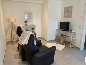 Uma sala de estar moderna com um sofá de couro preto, uma luminária de chão, uma televisão sobre um móvel de madeira e uma pequena estante com objetos decorativos.