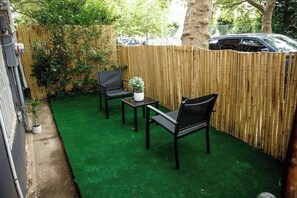 Een klein terras buiten, omringd door een bamboescherm, met twee stoelen, een klein tafeltje en een bloempot.