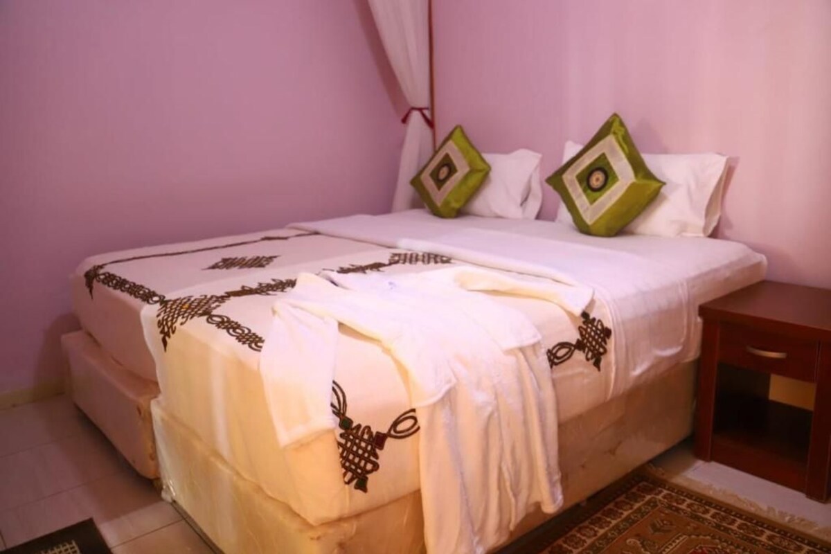 Una cama con una colcha blanca, almohadas decorativas, una mesita de noche de madera y una alfombra estampada en el suelo.