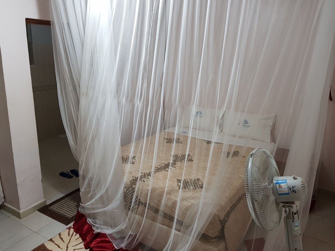 A bedroom with a bed, a standing fan, and a sheer curtain.