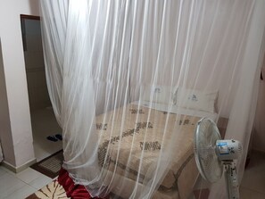A bedroom with a bed, a standing fan, and a sheer curtain.