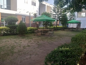 Un área de estar al aire libre con mesas y sillas bajo sombrillas verdes, rodeada de setos cuidados y un edificio al fondo.