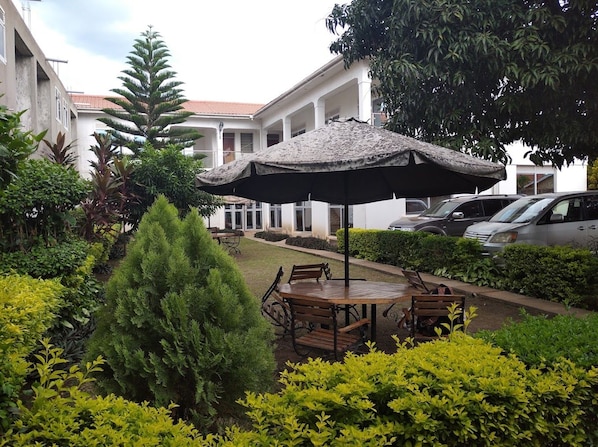 A courtyard with a round wooden table and chairs, surrounded by lush greenery and a large umbrella.