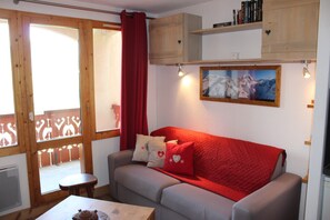 Ett mysigt vardagsrum med en grå soffa, röda kuddar, ett litet bord och ett fönster med träpersienner.