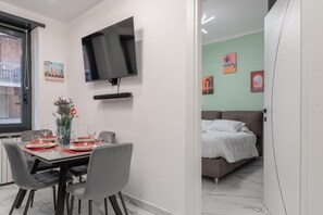 A modern living room with a flat-screen TV mounted on the wall, a dining area with a table set for two, and a bedroom with a bed and a nightstand. - Maison Tresor by Wonderful Italy (Turin)