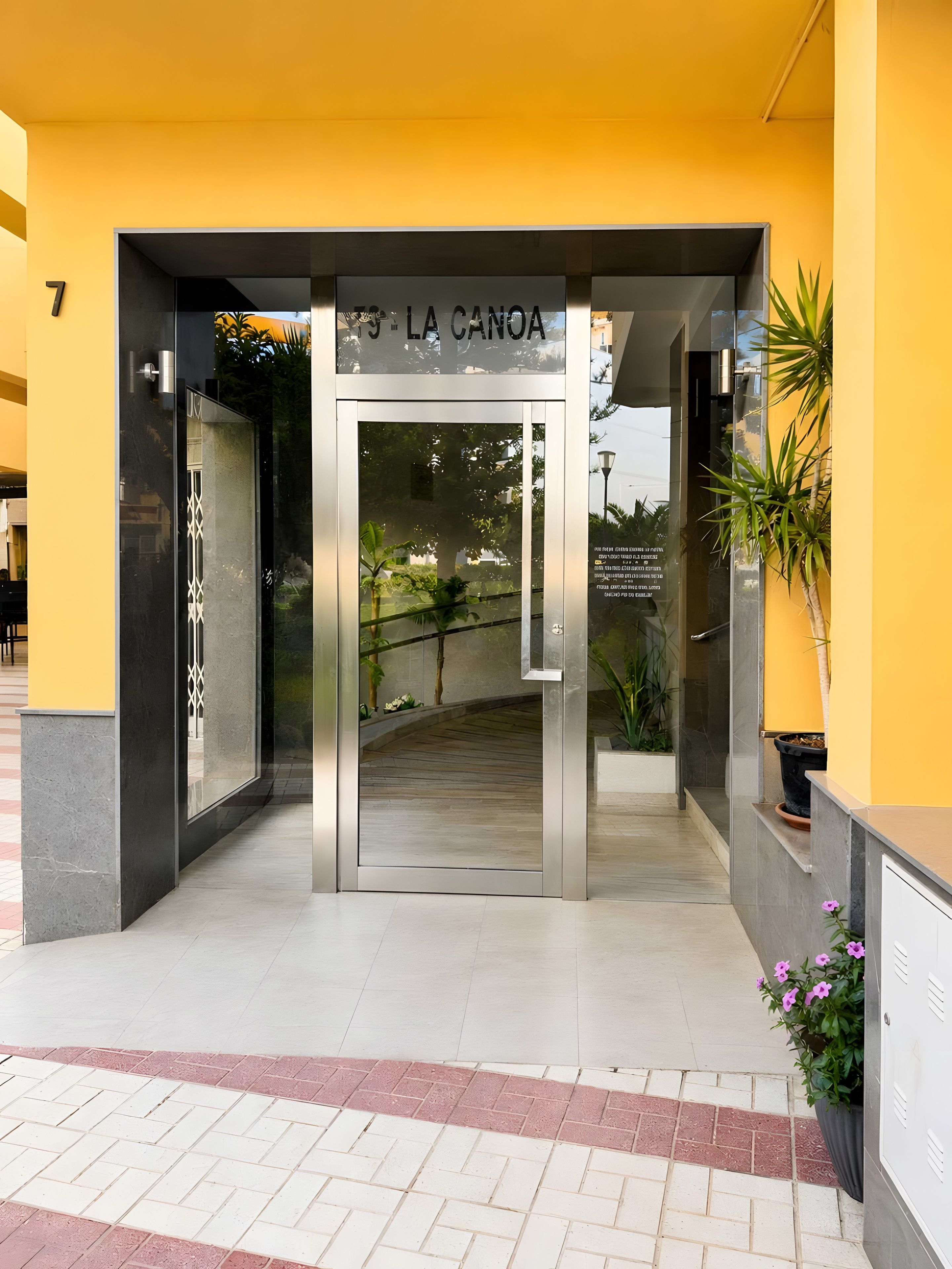 L’entrée d’un hôtel, avec une porte tournante en métal, une enseigne au-dessus et un pot de fleurs sur le côté droit.