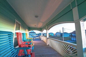 A covered porch with colorful Adirondack chairs and a view of houses in the background. - Our house is not fancy; it is welcoming. (Bolivar Peninsula)