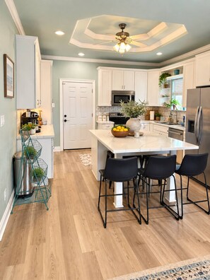 A modern kitchen with white cabinets, a central island with bar stools, a stainless steel refrigerator, and a ceiling fan. - Our house is not fancy; it is welcoming. (Bolivar Peninsula)