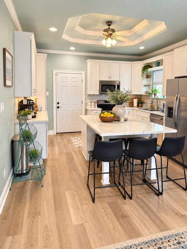 Una cocina moderna con armarios blancos, una isla central con taburetes de bar, un refrigerador de acero inoxidable y un ventilador de techo.
