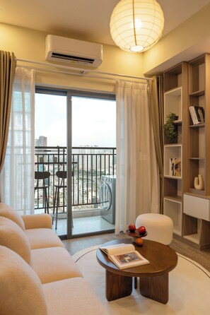 A modern living room with a sofa, a coffee table with books and apples, a bookshelf, and a balcony with a view of buildings. - 2BR Corner | King + Queen | Soho D1 | Floor 24 (Thành phố Hồ Chí Minh)