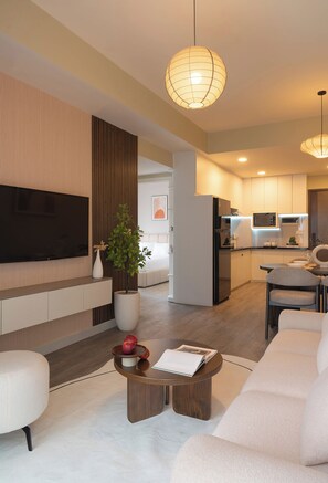 A modern living room with a flat-screen TV, a large potted plant, a coffee table with apples and a book, and a sofa. - 2BR Corner | King + Queen | Soho D1 | Floor 24 (Thành phố Hồ Chí Minh)