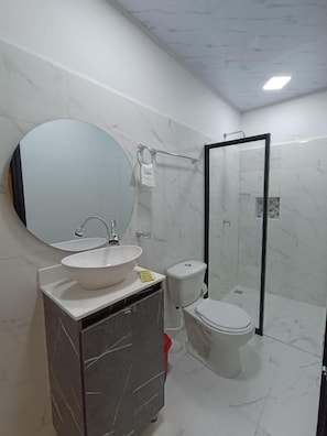 Un bany amb un mirall rodó, un lavabo, un vàter i plat de dutxa amb envàs de vidre.
