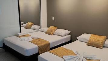 A hotel room with three beds, each with a white mattress and a brown headboard. There is a large mirror on one wall and a ceiling fan.