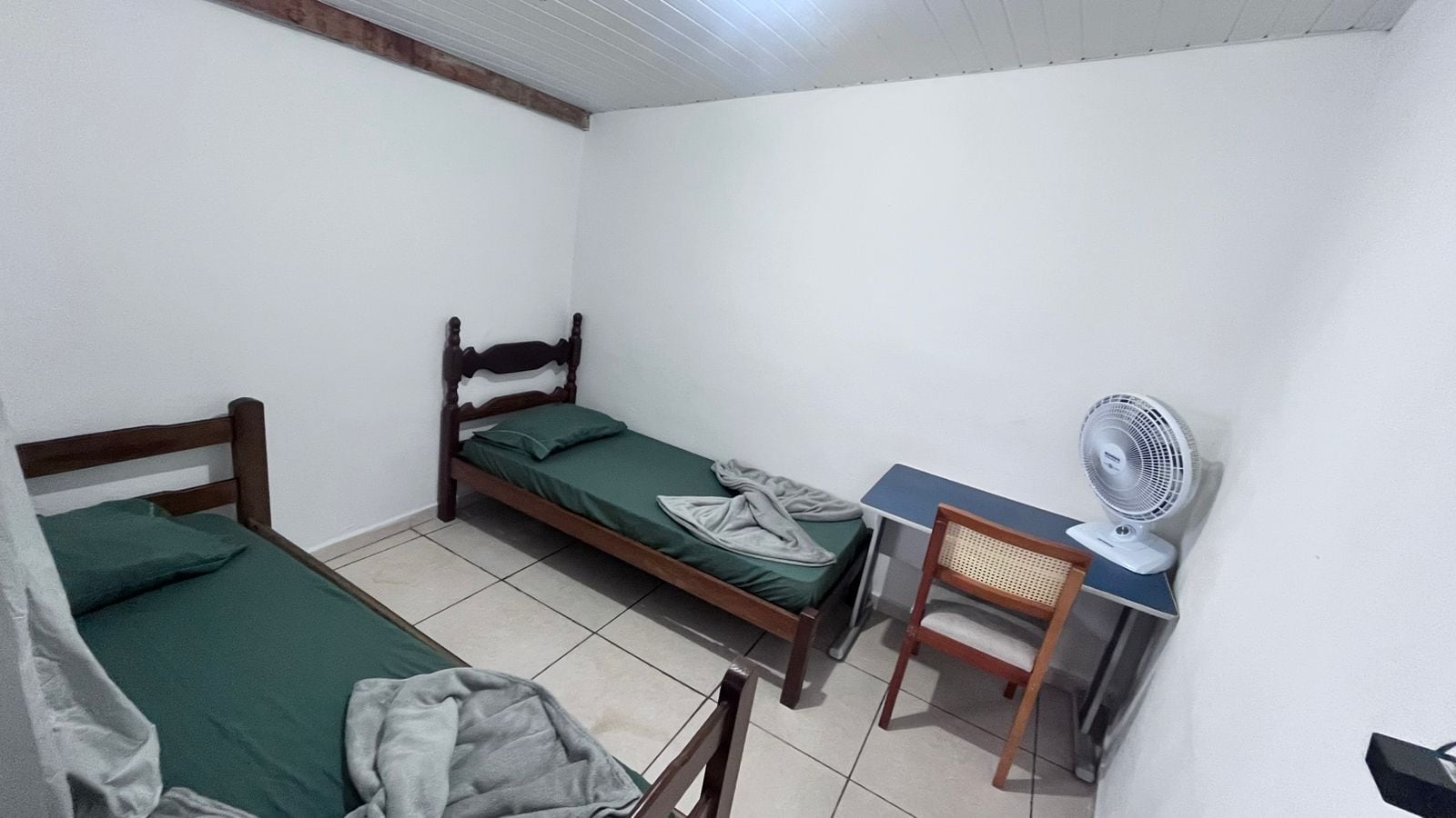A small, single-room accommodation with two wooden beds, a desk, a chair, and a standing fan.