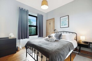 A bedroom with a bed, a bedside table with a lamp, a window with curtains, a door, and a framed picture on the wall. - Serene 2BR Stay in Penrith | Sleeps 4+Parking+WiFi (Penrith)
