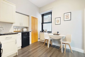A compact kitchen with white cabinetry, a black dishwasher, a wooden door, and a dining area with a white table and chairs. - Serene 2BR Stay in Penrith | Sleeps 4+Parking+WiFi (Penrith)