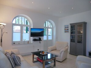 Ein Wohnzimmer mit Flachbildfernseher an der Wand, einem Glastisch, zwei weißen Ohrensesseln, einer Couch und einer Vitrine mit Glastüren.