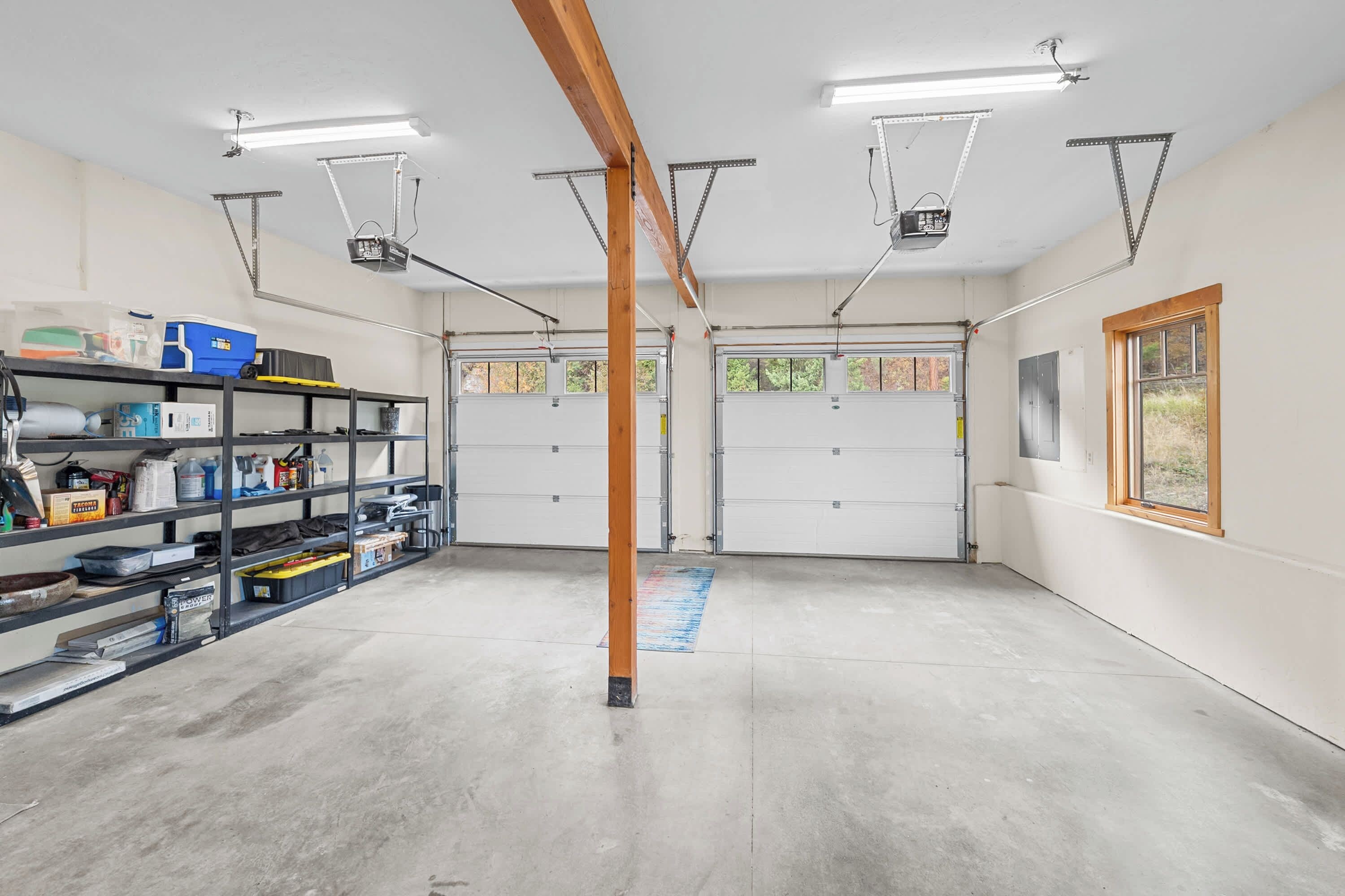 Een garage met twee witte garage deuren, een houten balk, fluorescent verlichting en een plank met diverse spullen.