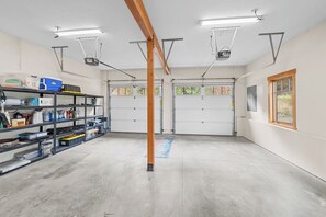 A garage with two white garage doors, a wooden beam, fluorescent lighting, and a shelf with various items.