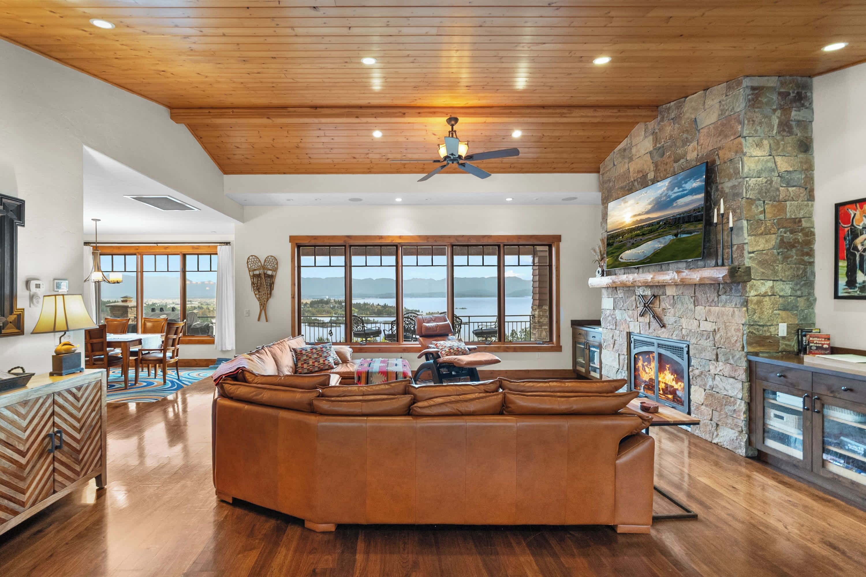 A spacious living room with a large brown leather sectional sofa, a stone fireplace, a mounted TV, and a view of the outdoors through large windows.