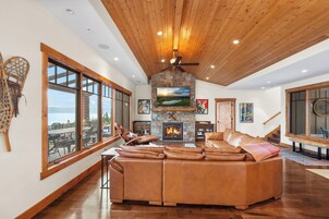 Un salón amplio con chimenea de piedra, sofá de cuero tipo seccional, ventanas grandes y ventilador de techo.