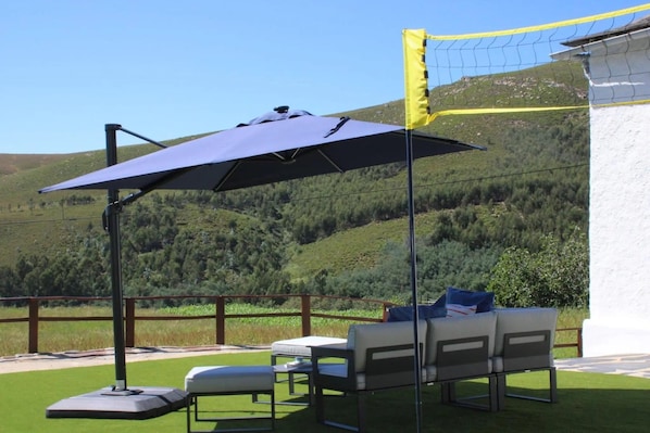 An outdoor patio with a table and chairs, a large umbrella, and a view of a mountainous landscape.