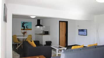 Una sala de estar con sofá, una mesa de centro con un tablero de ajedrez, un comedor con sillas, una cocina con microondas y un televisor montado en la pared.